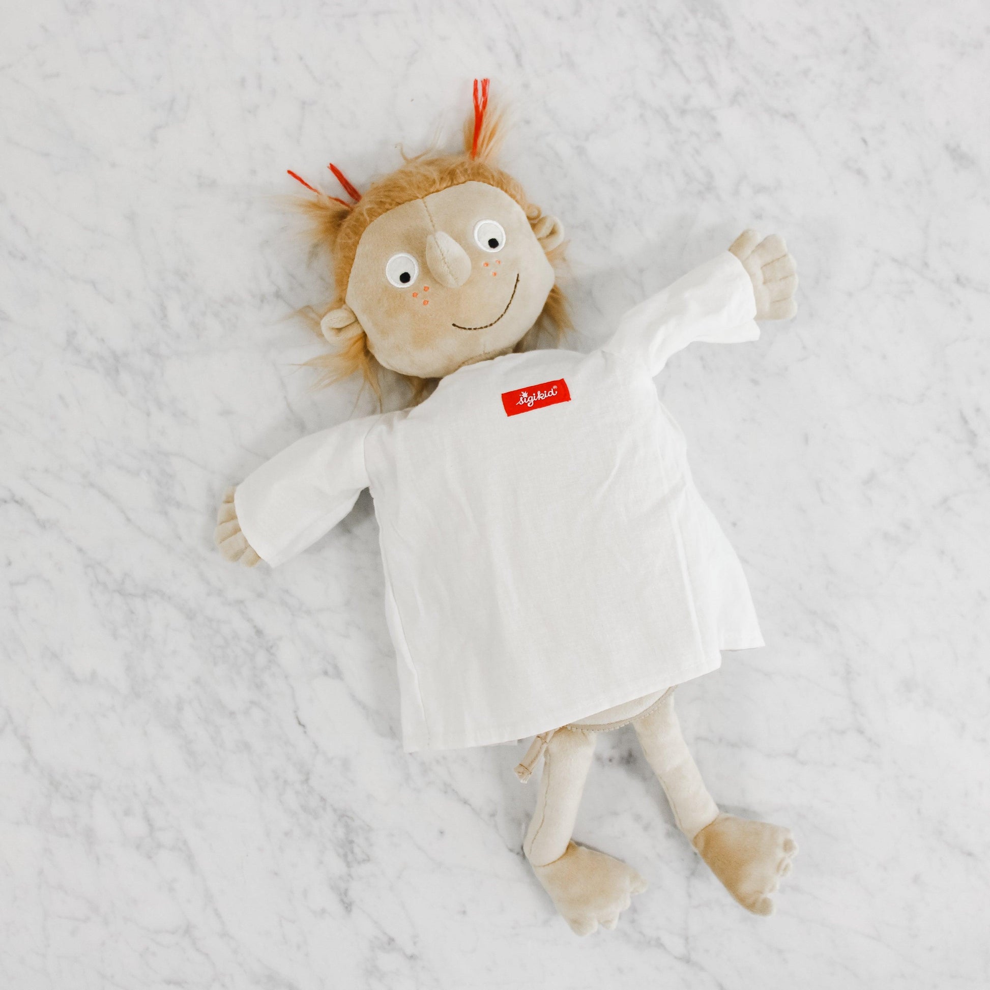 The Sigikid - Rosi the Little Patient plush doll by Grace & Company ltd lies on a white marble surface.