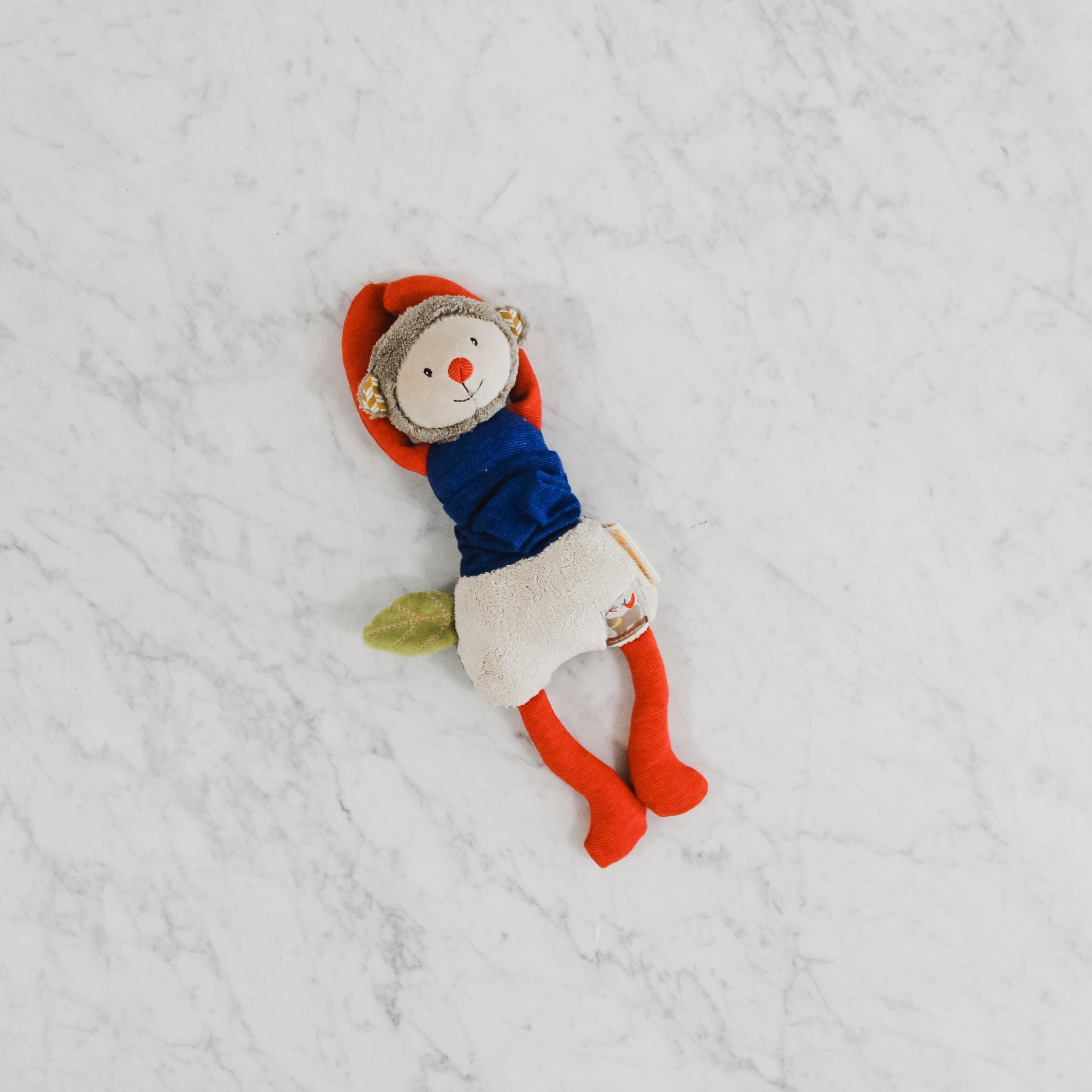 The Moulin Roty - Monkey by Grace & Company ltd, with a red hat and blue shirt, rests on white marble.