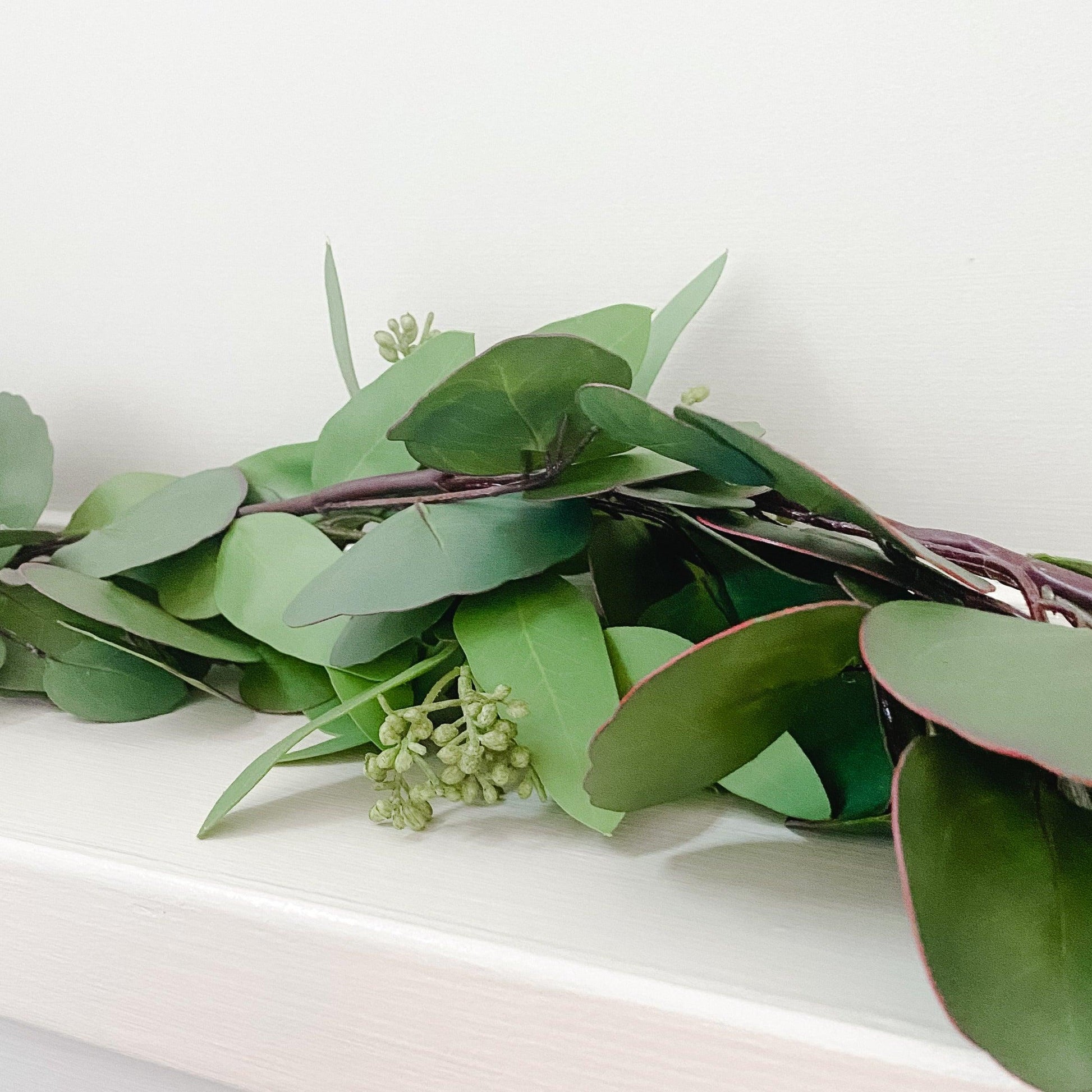The Grace & Company Eucalyptus Garland with small buds rests on a white surface against a pale background.
