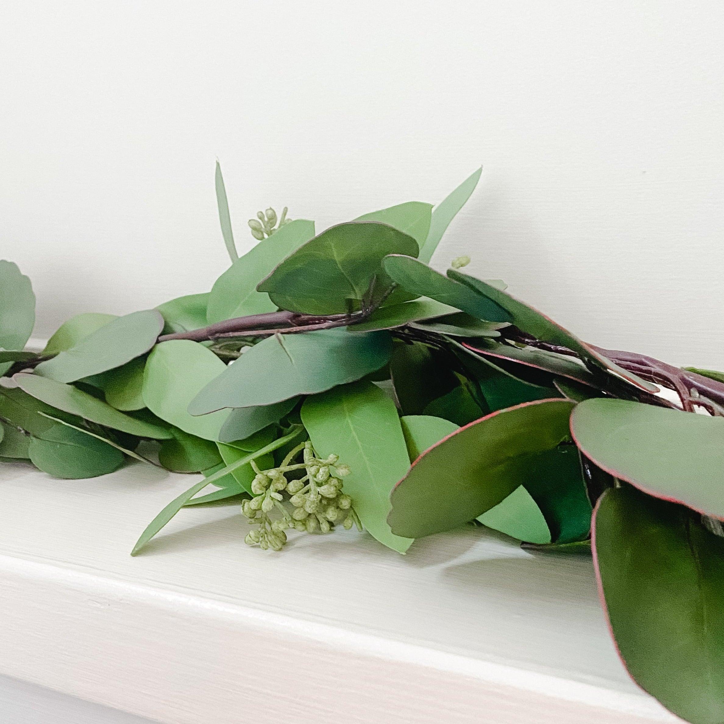 The Grace & Company Eucalyptus Garland with small buds rests on a white surface against a pale background.