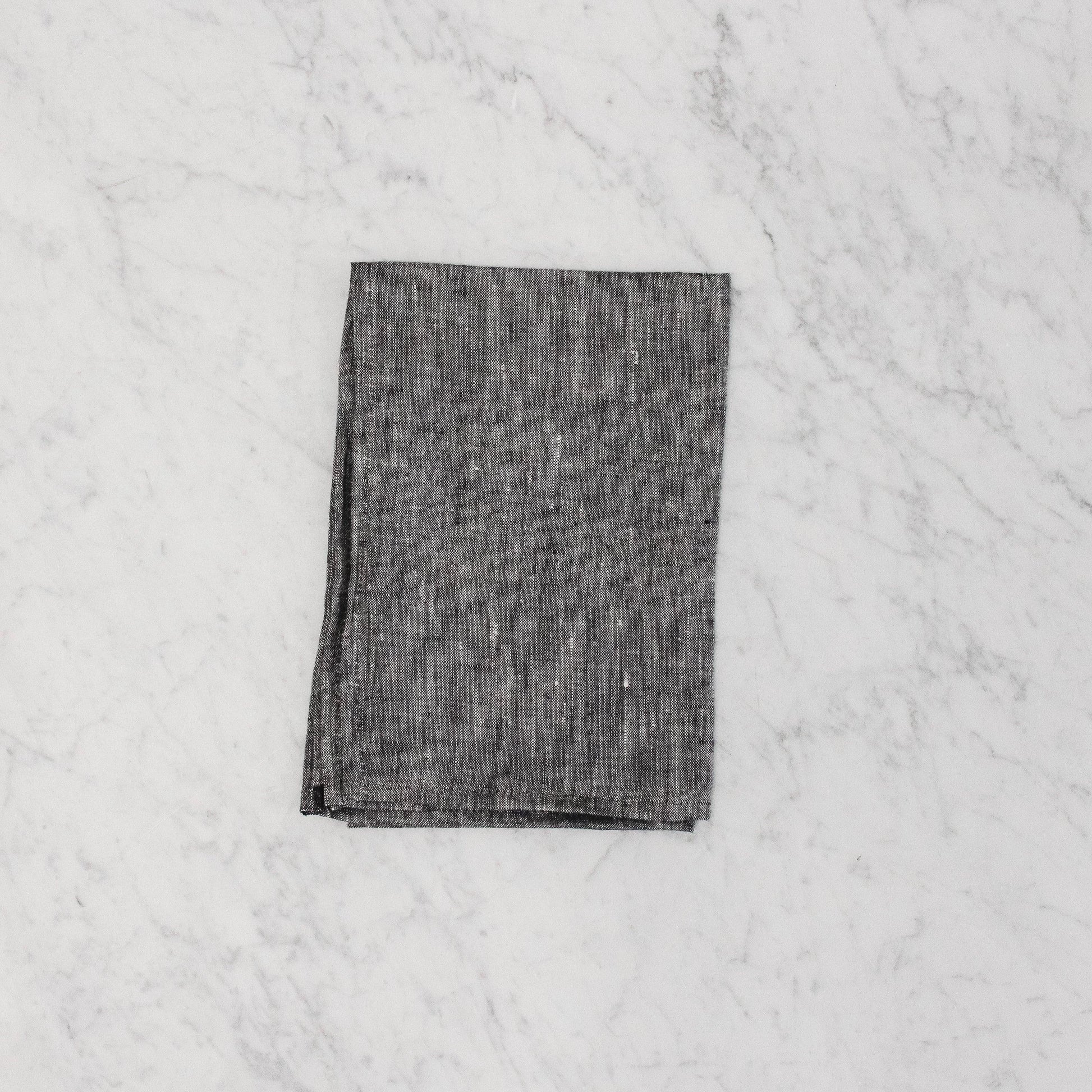 A G&C - Tea Towel by Grace & Company ltd, folded dark gray, on a white marble surface.