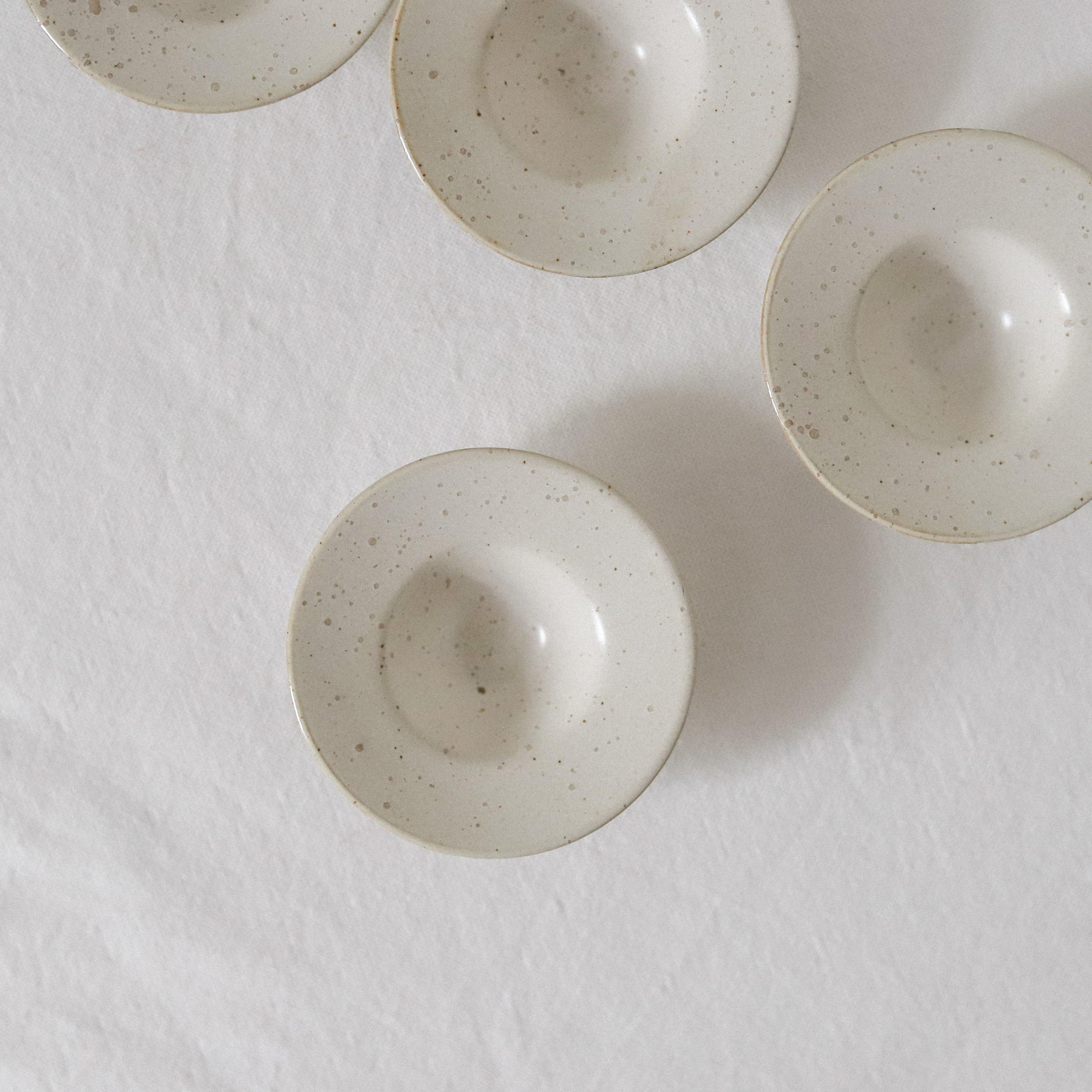 Two white, speckled ceramic Pion tiny bowls by Grace & Company are displayed on a white cloth in soft natural light.