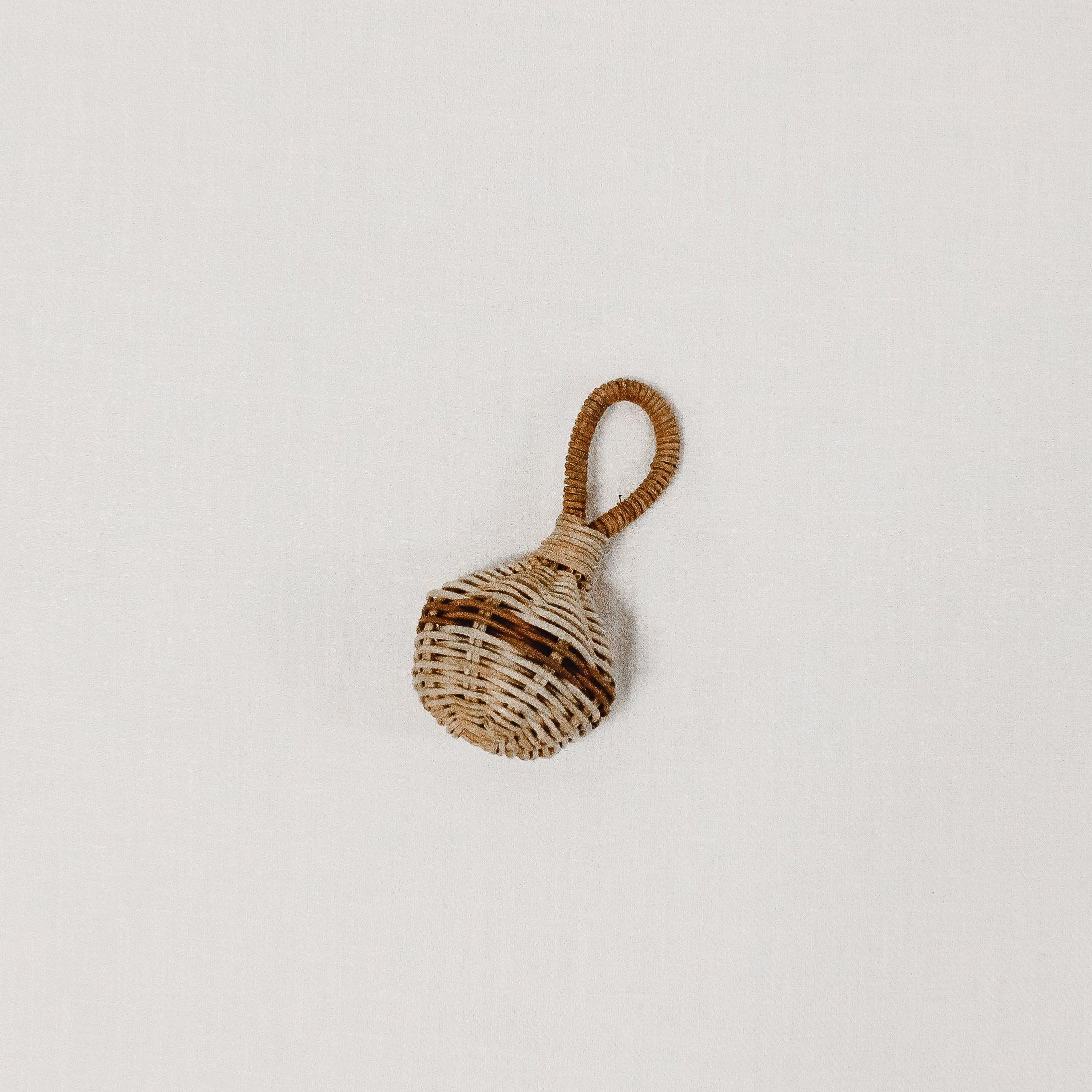 The Grace & Company Olli Ella Raya Rattle is a small, woven rattle with a looped handle on a white background.