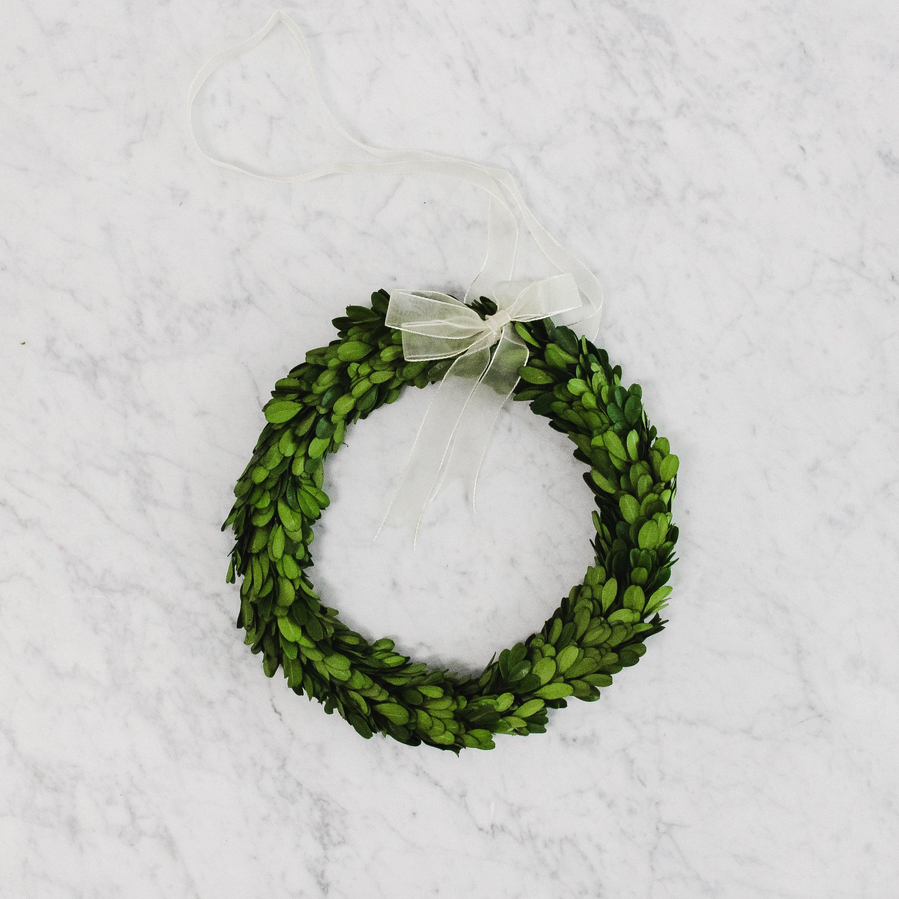 The Grace & Company Boxwood Wreath with a White Ribbon is displayed on a white marble background.
