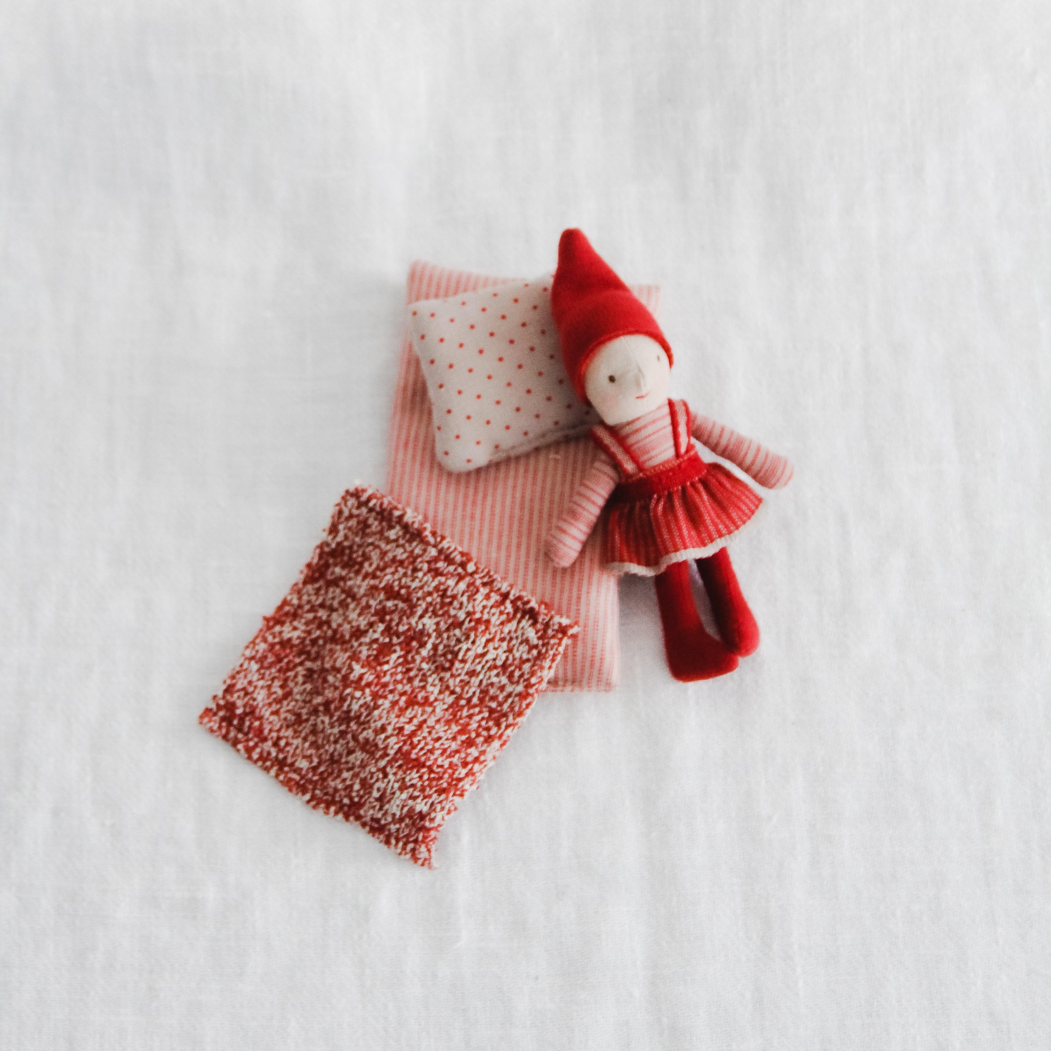 Maileg - Pixy Elfie in a Box by Grace & Company: a small elf doll in red rests on pillows and a blanket.