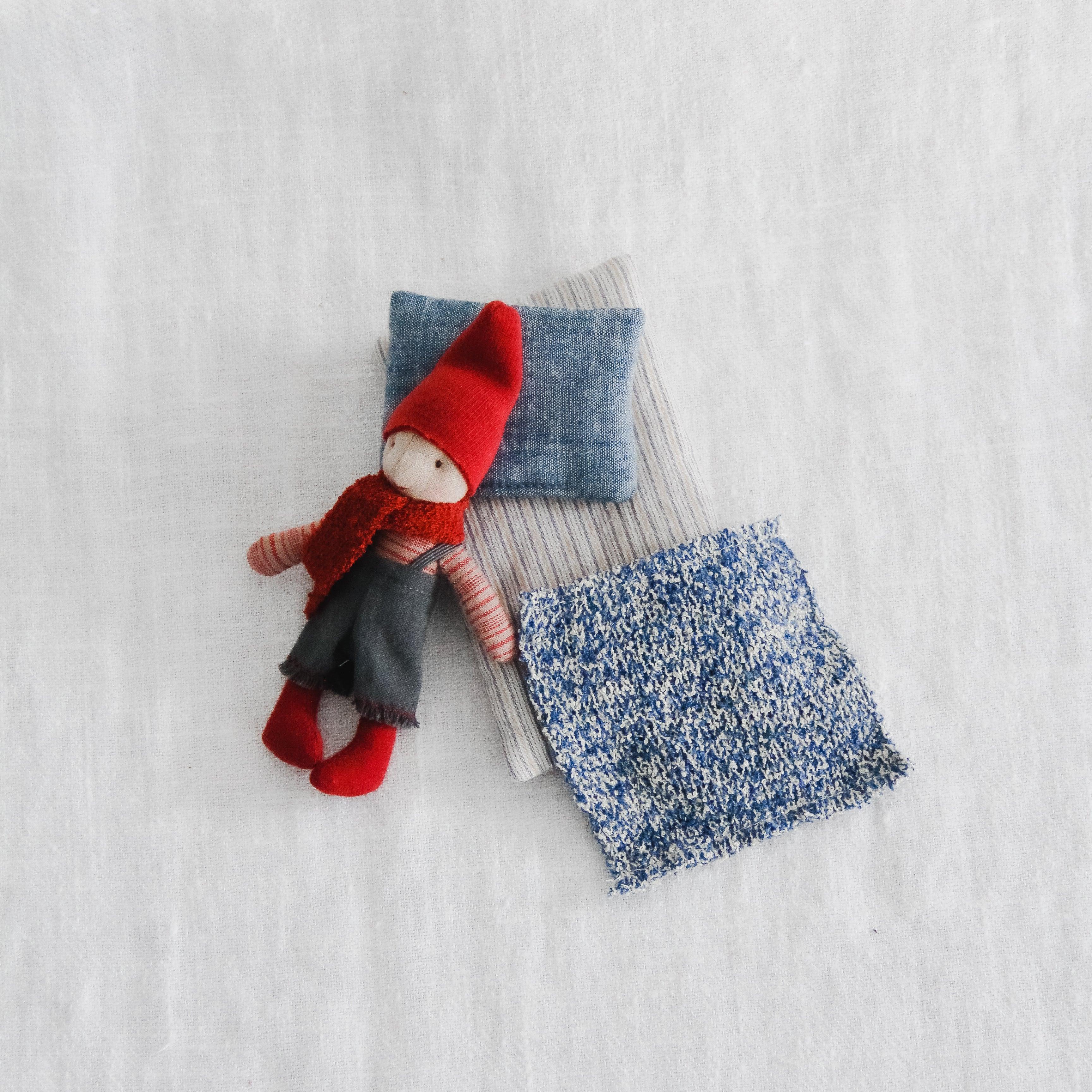 The Maileg - Pixy Elf in a Box by Grace & Company rests on fabric pillows and a blue blanket on a white surface.