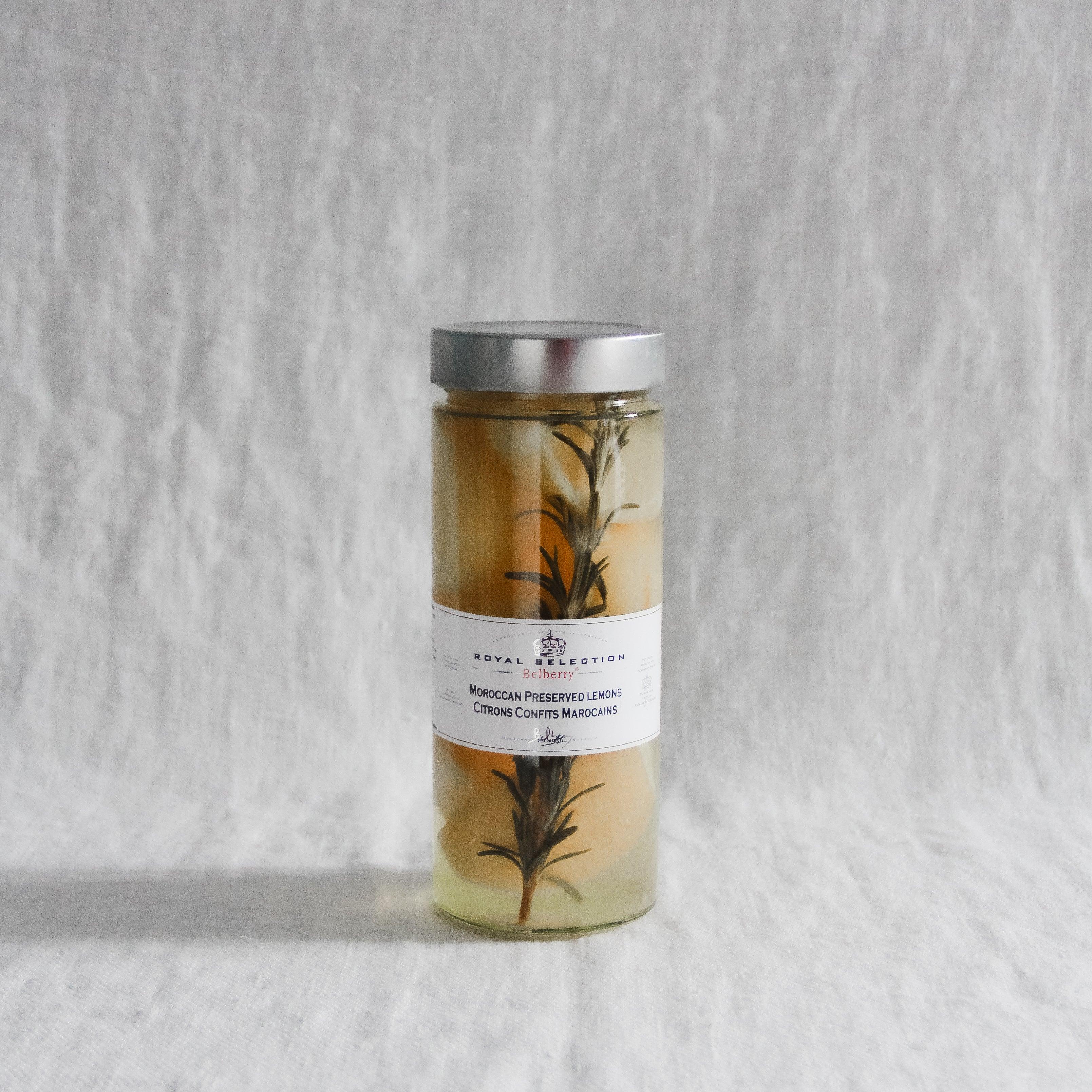 A glass jar with a silver lid, labeled Grace & Company Royal Selection Moroccan Preserved Lemons, with lemons and herbs.