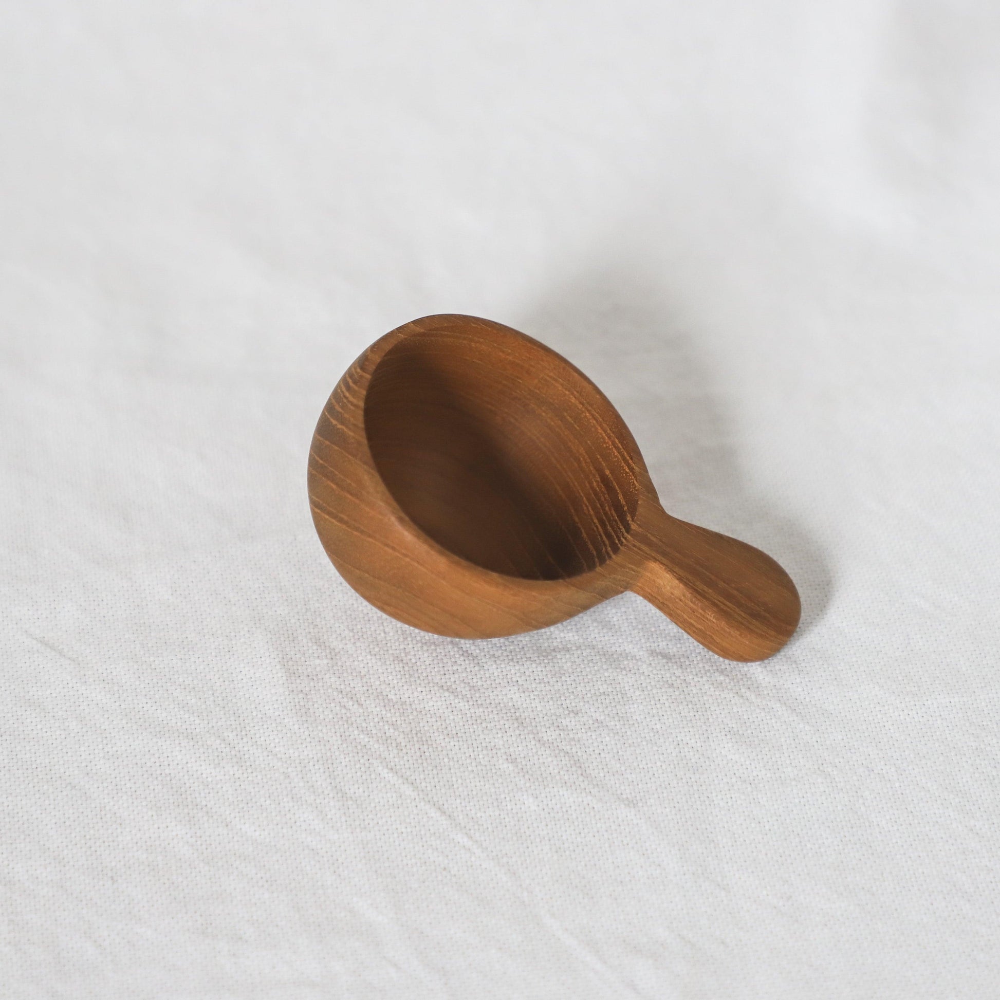 The Grace & Company Teak Spoon with a short handle rests on a white textured fabric background.