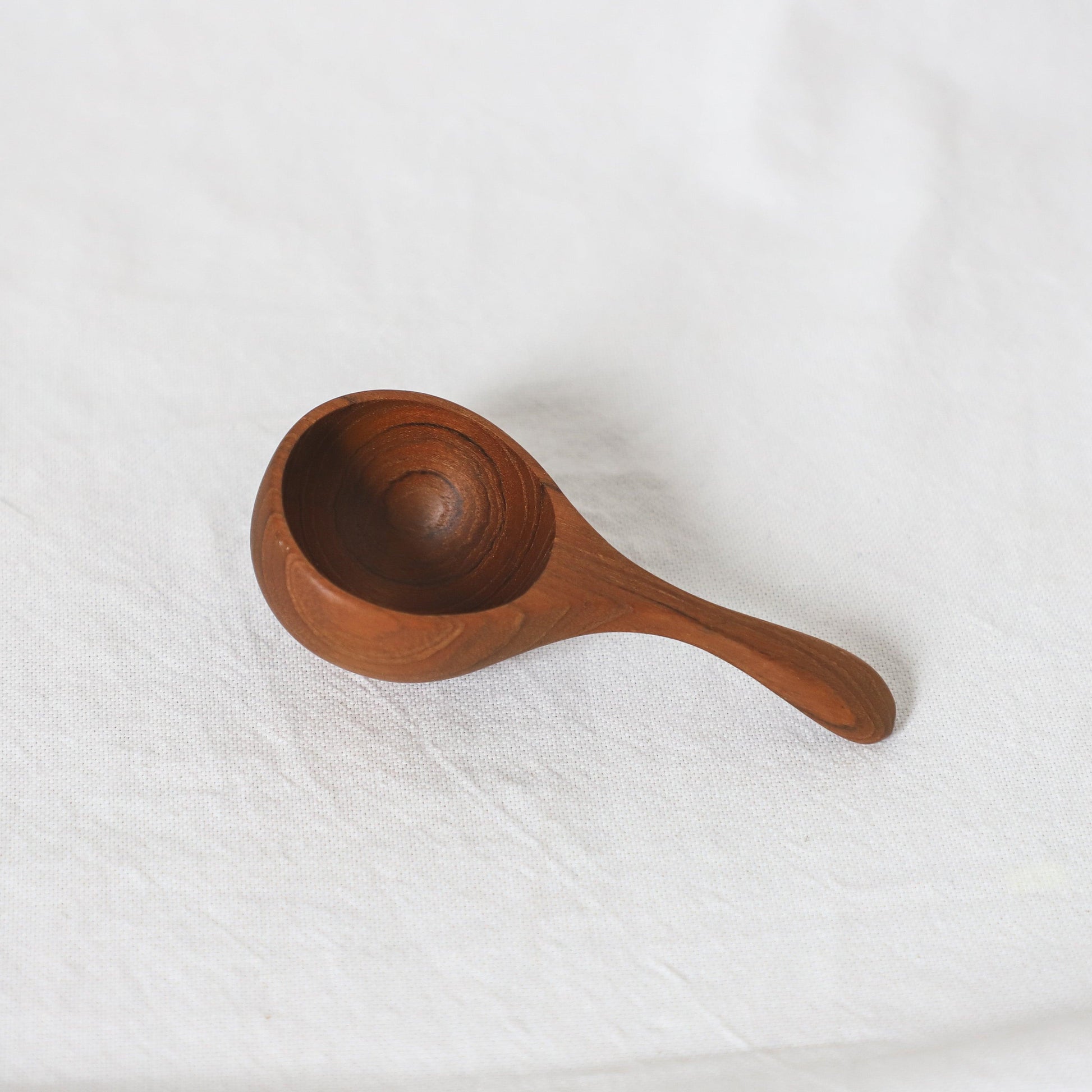The Grace & Company Teak Spoon features a round bowl and curved handle, shown on a white fabric background.