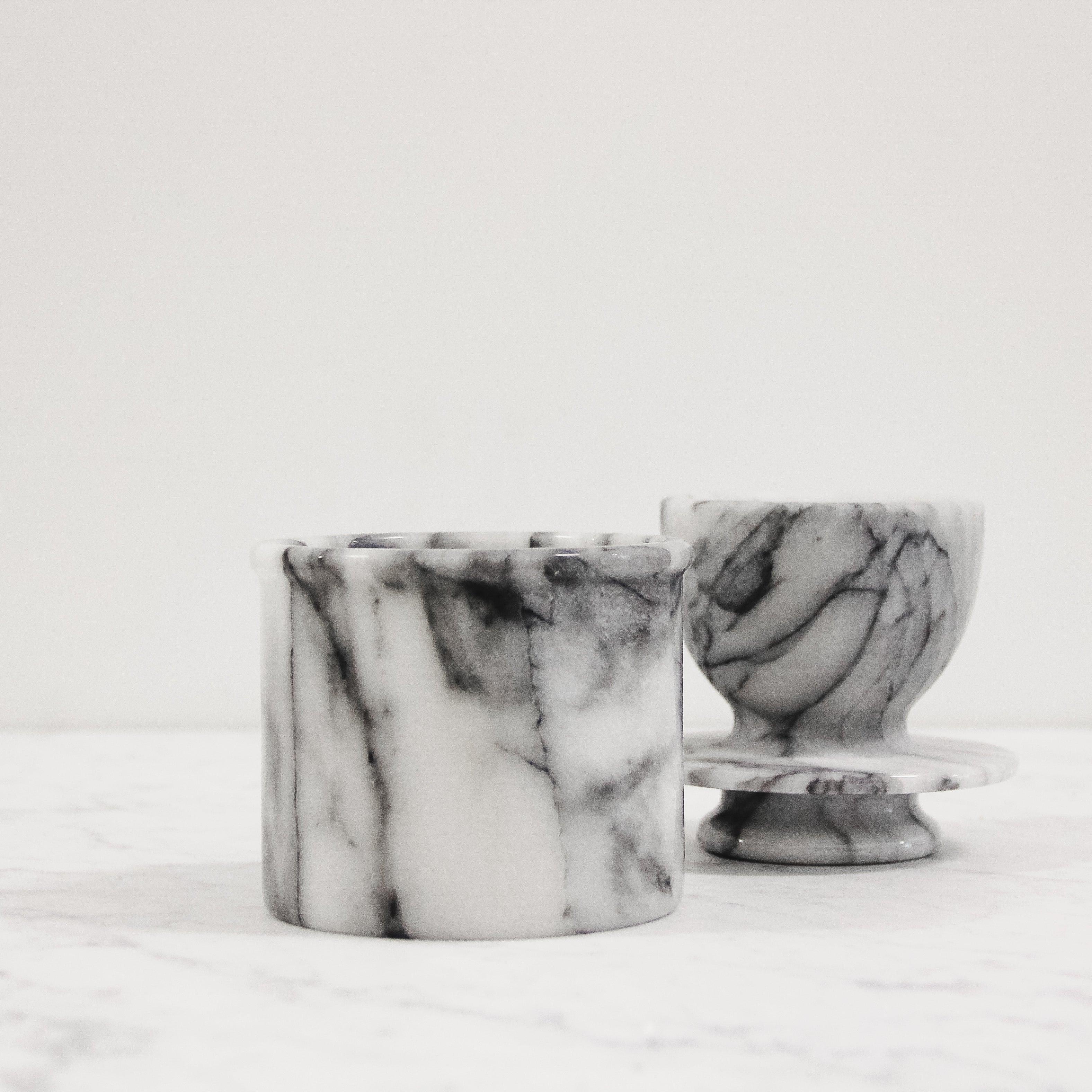 Two Grace & Company ltd Marble Butter Bells with gray and white veining on a white marble surface and background.