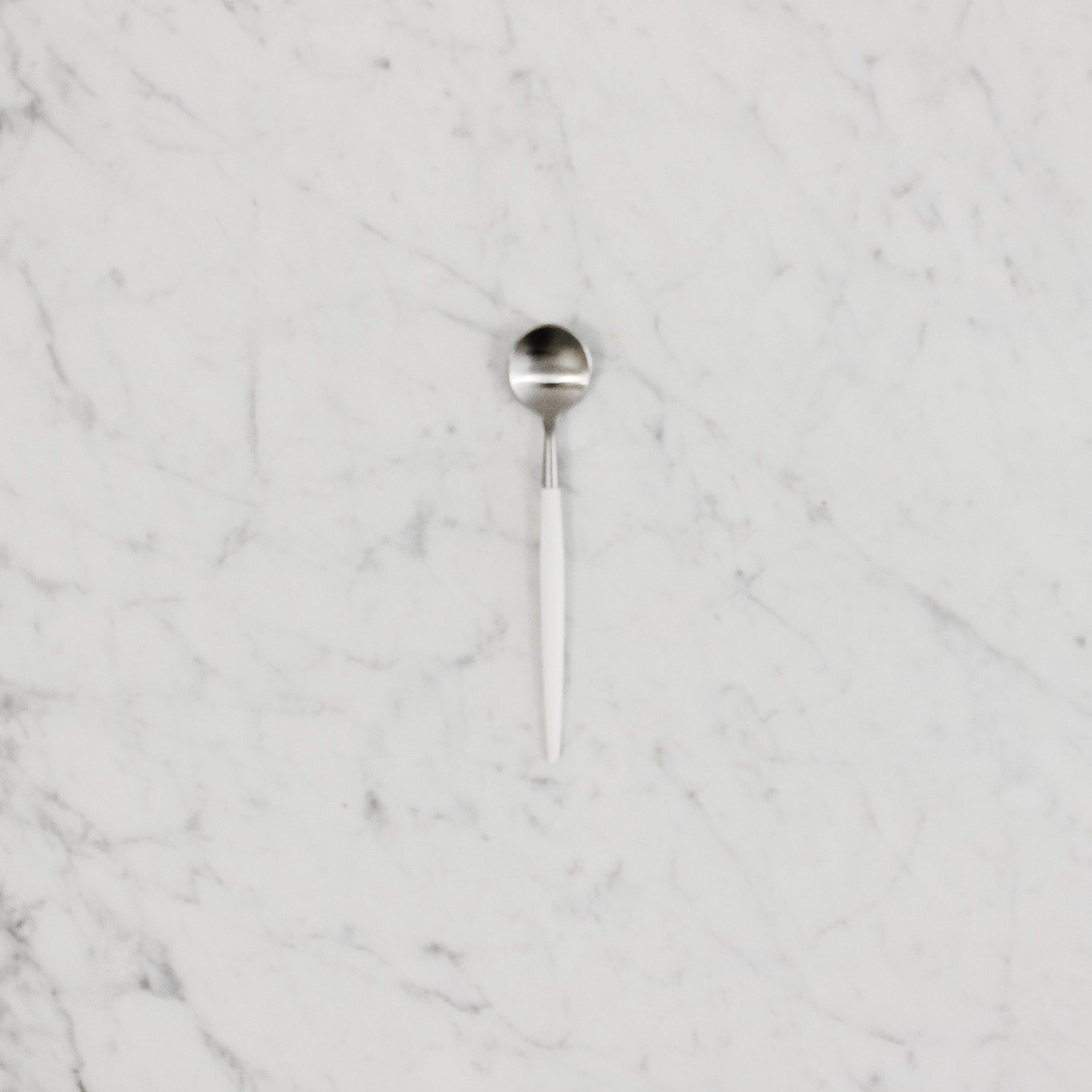 The Cutipol Goa Moka Spoon by Grace & Company ltd with a white handle rests on a white marble surface.