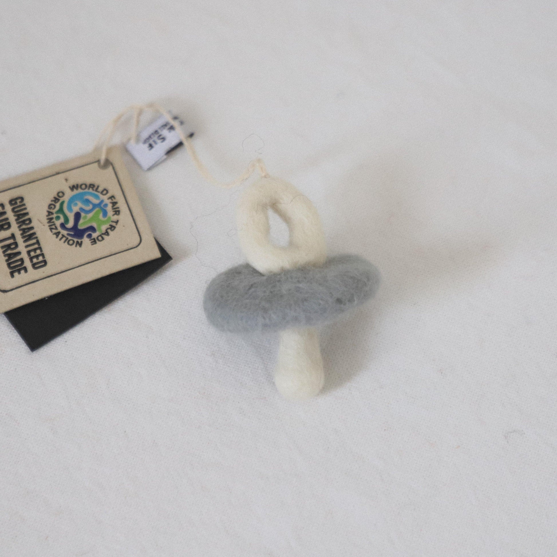 The Grace & Company Scandi-Felted Pacifier features a gray ring, white handle, and fair trade tag on a white surface.