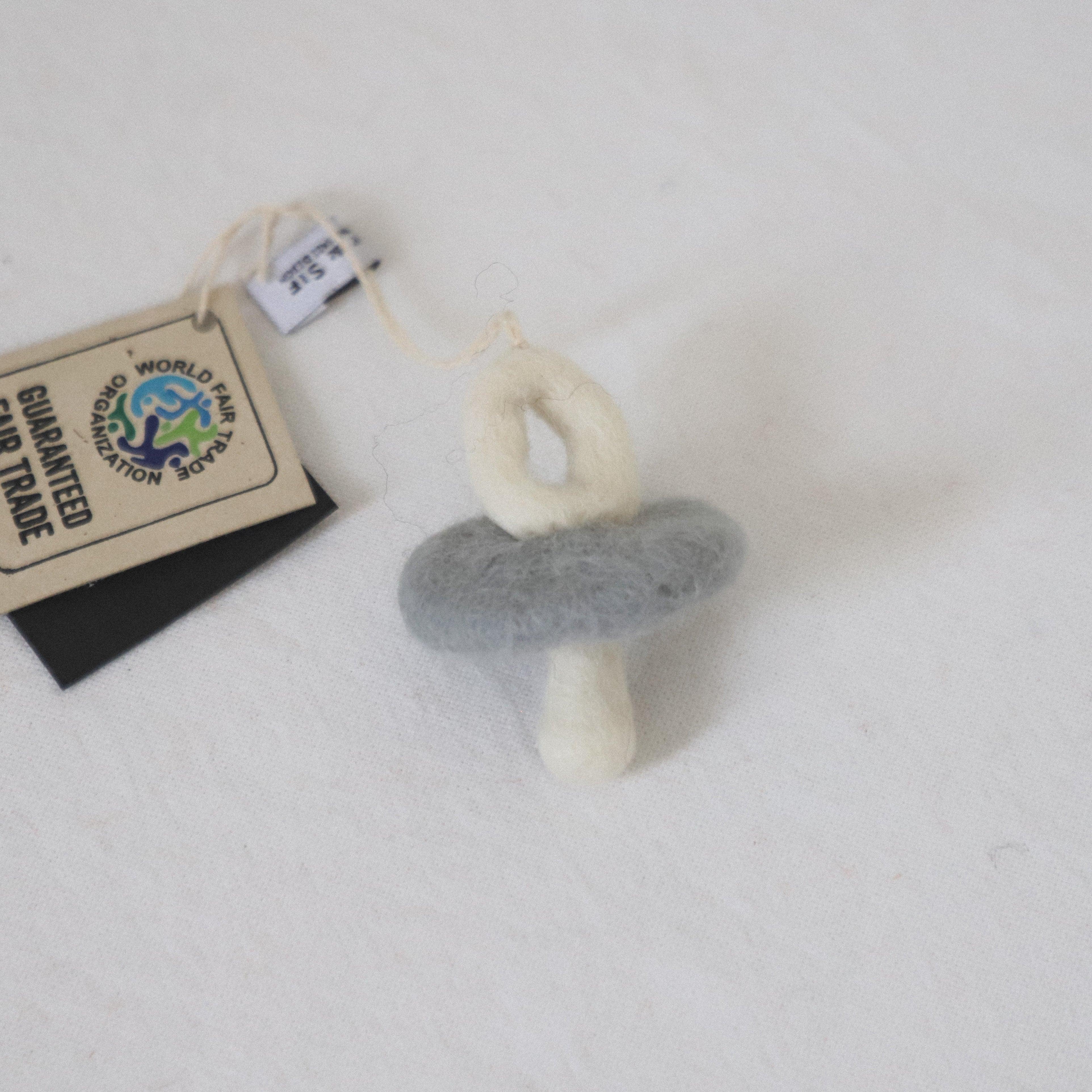 The Grace & Company Scandi-Felted Pacifier features a gray ring, white handle, and fair trade tag on a white surface.