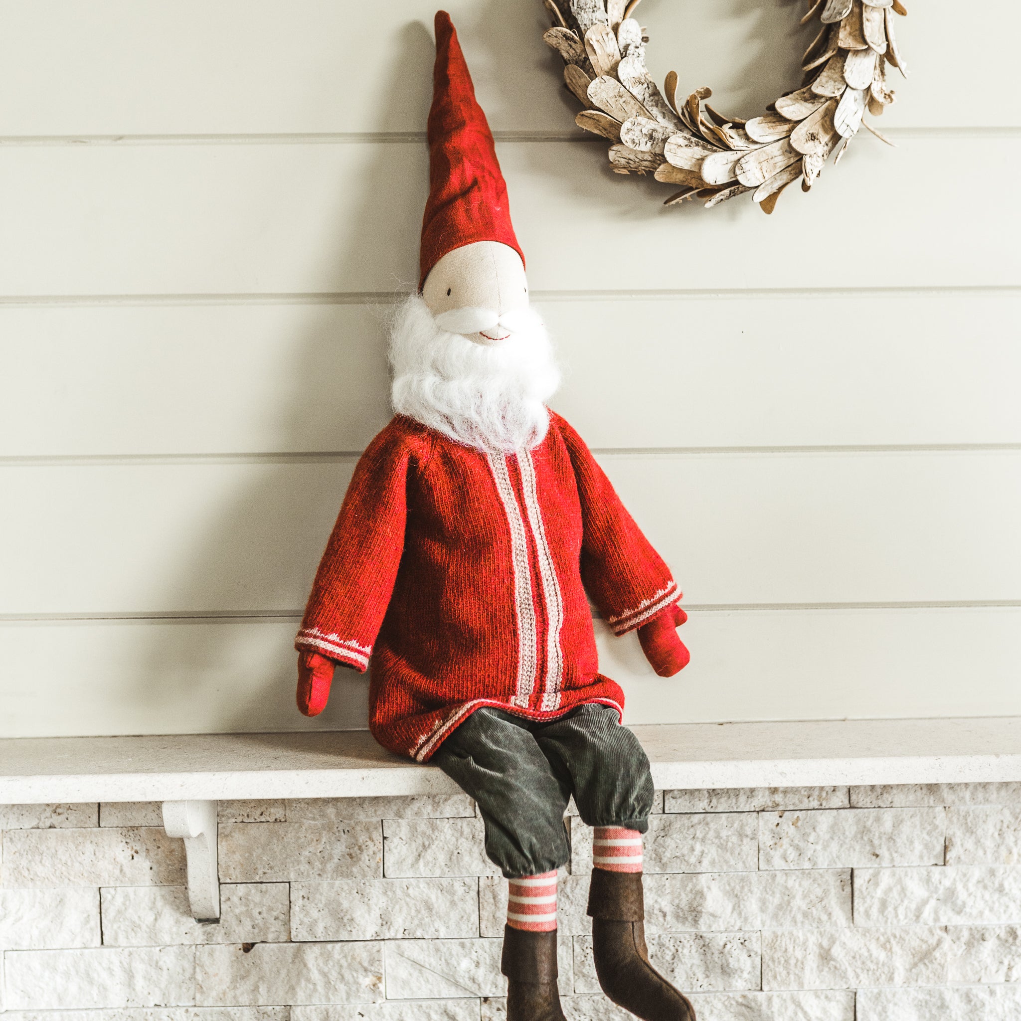 A Maileg - Santa plush doll by Grace & Company in red sits on a shelf under a wreath on a light wall.